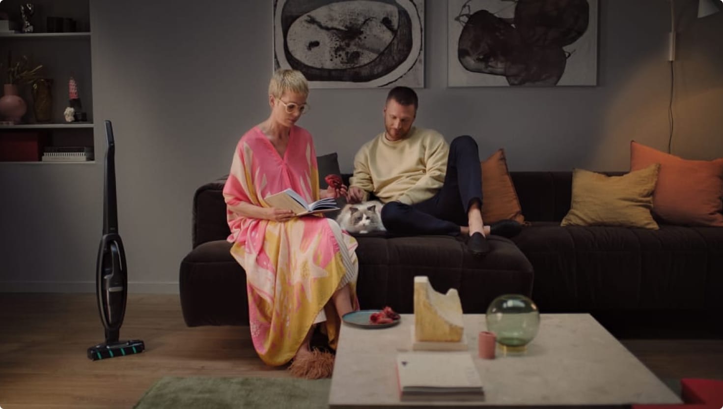 Man and a woman sitting on a couch in a living room with a cat in between them.